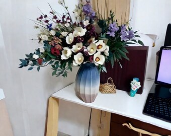 Op de afbeelding: Een witte keramische vaas met blauwe en grijze ombre-glazuur bevat een boeket witte, paarse en rode bloemen. De vaas staat op een witte tafel met een kleine rieten mand en een tealblauwe vaas met witte bloemen.