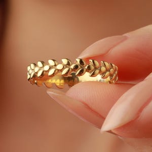 May include: A gold ring featuring a repeating floral design. The ring is held between fingers, showcasing its intricate details and polished finish. The ring's design is a continuous pattern of small, stylized leaves.
