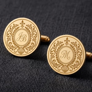 Op de afbeelding: Een paar ronde goudkleurige manchetknopen met een ingewikkelde gegraveerde filigraanrand en een centraal rond embleem. De ene manchetknop toont de cursieve initialen JB, terwijl de andere manchetknop de initiaal M in het midden weergeeft.