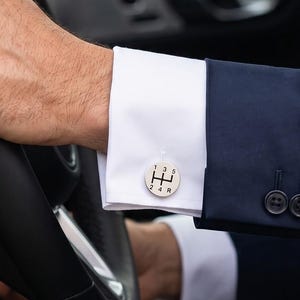 May include: A round silver-toned cufflink featuring a black manual transmission gear shift pattern with numbers 1, 3, 5 and 2, 4, R printed on the face. The cufflink is secured on the white cuff of a long-sleeved shirt worn under a navy blue blazer.