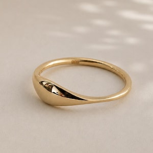 May include: A polished gold ring featuring a smooth, rounded, and slightly tapered band that widens into a solid, teardrop-shaped focal point. The metal surface is reflective and captures light, showcasing a clean, continuous, and seamless design.