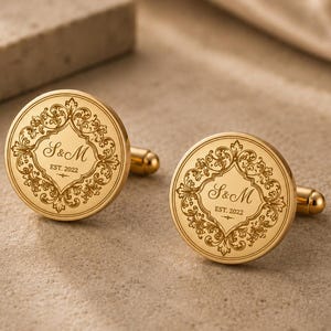 May include: A pair of round gold-toned cufflinks featuring an intricate engraved filigree border surrounding the central text S&M and EST. 2022. The cufflinks have a polished metallic finish and a standard T-bar fastening mechanism on the back.