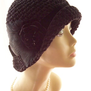 May include: A dark brown cloche hat with a textured knit design. The hat features a decorative black flower and feathers on the side. A black band encircles the hat, and the brim is slightly turned up. The hat is displayed on a mannequin head.