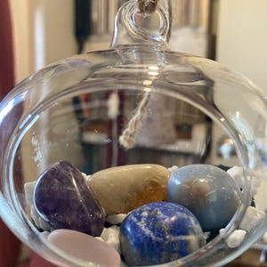 Puede incluir: Una bola de cristal con un lazo de cuerda en la parte superior, que contiene seis piedras pulidas: amatista, citrino, ágata azul, lapislázuli, cuarzo rosa y cuarzo transparente. Las piedras están anidadas en roca decorativa blanca.