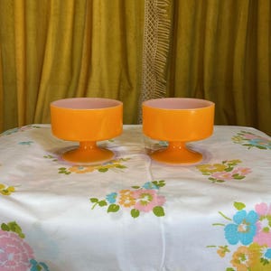 May include: Two orange glass dessert bowls with a pink interior. The bowls have a pedestal base and a cylindrical body. They are set on a white tablecloth with a floral pattern. The background features a yellow curtain.