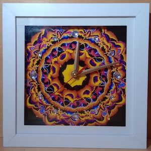 Può includere: Orologio da parete quadrato con cornice bianca e design astratto colorato. Il quadrante presenta un motivo vibrante di tonalità gialle, arancioni, viola e blu, con accenti di cristallo. Le lancette dell'orologio sono dorate e rosse.