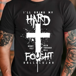 Może przedstawiać: Czarna koszulka z białym krzyżem i napisem "I'LL BRING MY HARD FOUGHT HALLELUJAH". Koszulka zawiera również słowa "HEART FELT" i "BEEN THROUGH HELL".