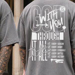May include: A charcoal gray short-sleeve t-shirt featuring white graphic text. The front displays "GOD IS WITH YOU". The back features "GOD IS WITH YOU THROUGH IT ALL" with a checklist including "EVERY SEASON", "EVERY DOUBT", "EVERY STRUGGLE", "EVERY BATTLE", and "EVERY VICTORY".