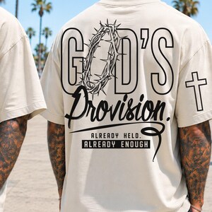 May include: A beige t-shirt featuring a black graphic design of a crown of thorns and the text "GOD'S Provision" on the front. The back displays the same crown of thorns, the text "GOD'S Provision", a cross, and the words "ALREADY HELD. ALREADY ENOUGH."