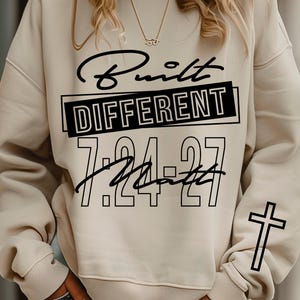 Pode incluir: Camisola creme com o texto "Built Different" em escrita preta e letras de bloco. Os números "7:24-27" e a palavra "Matt" também estão impressos na frente. Uma cruz preta está na manga direita.