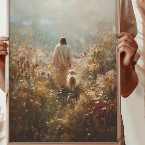 May include: A framed print shows a figure in a white robe walking through a field of wildflowers, with a lamb. The artwork has a soft, painterly style with a warm colour palette of greens, yellows, and pinks. The frame is a light brown colour.