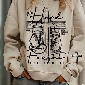 May include: Cream-colored sweatshirt with a graphic design featuring boxing gloves on a cross, with the words "I'll bring my heart felt fought hallelujah" and "been through hell". A small cross is on the sleeve.