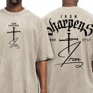 Pode incluir: Camiseta bege com uma cruz preta e as palavras "Iron Sharpens Iron" impressas na frente e atrás. A parte de trás também inclui "Prov. 27:17". A camiseta tem um visual vintage.