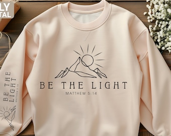 May include: A beige sweatshirt with a black line drawing of a mountain range and sun with the text "BE THE LIGHT" and "MATTHEW 5:14" below the image.