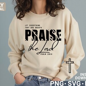 May include: A beige sweatshirt with black text that reads "Let everything that has breath praise the Lord Psalm 150:6". The word "praise" is in a bold, striped font. A white outline of a cross is on the right sleeve of the sweatshirt.