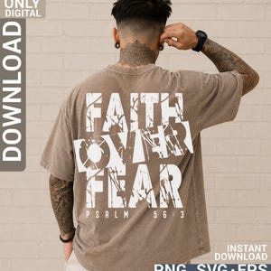 May include: A brown short-sleeved t-shirt featuring a large white graphic on the back with the text FAITH OVER FEAR and PSALM 56:3. The text has a distressed, paint-splatter effect. The shirt has a relaxed fit and a faded, vintage-style finish.