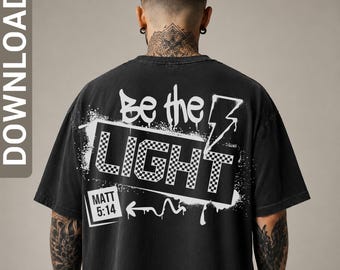 May include: A black short-sleeved t-shirt featuring a white graphic on the back. The design includes the text "Be the LIGHT" in a graffiti style with a checkered pattern inside the letters, a lightning bolt, and a box containing "MATT 5:14".