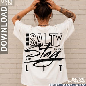 May include: An off-white short-sleeve t-shirt featuring a black graphic print on the back. The text reads BE SALTY STAY LIT with the reference MATT 5:13-14. The design combines bold block lettering with a large, stylized brushstroke graphic element.