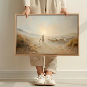 Op de afbeelding: Een ingelijste kunstdruk toont een figuur met een lam die een kudde schapen over een pad leidt. Het kunstwerk gebruikt een warm kleurenpalet met een heldere lichtbron. De lijst is gemaakt van lichtgekleurd hout.