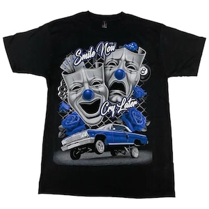 May include: Black t-shirt featuring a graphic design with the text "Smile Now Cry Later". The design includes comedy and tragedy masks with blue clown noses, blue roses, a lowrider car, and an 8-ball.