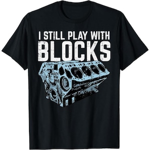 May include: Black t-shirt with the text "I STILL PLAY WITH BLOCKS" in white block letters. Below the text is a blue and gray illustration of an engine block. The shirt is made of a soft material.