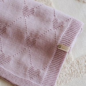 May include: A soft, pink knitted baby blanket with a heart pattern. The blanket has a small brown leather tag with the word "Little" on it.