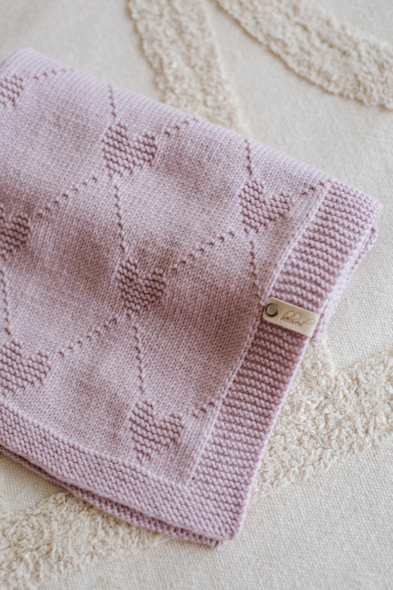 May include: A soft, pink knitted baby blanket with a heart pattern. The blanket has a small brown leather tag with the word "Little" on it.