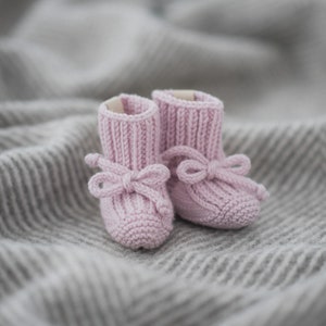 May include: A pair of pink knitted baby booties with bows. The booties are on a gray and white striped blanket.