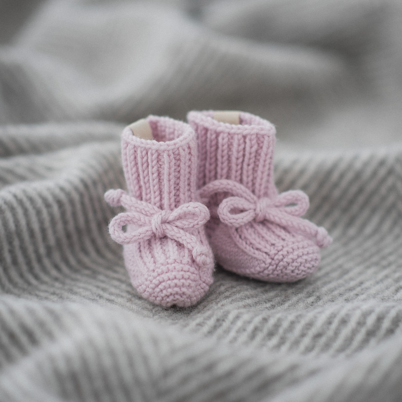 May include: A pair of pink knitted baby booties with bows. The booties are on a gray and white striped blanket.