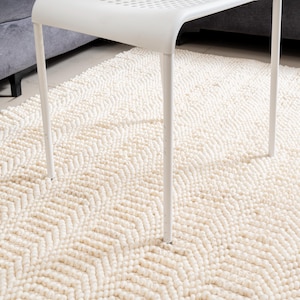 May include: Cream-colored, textured rug with a geometric chevron pattern. The rug is made of looped yarn, creating a soft, plush appearance. A white metal chair with a slatted seat and four legs is partially visible, resting on the rug.