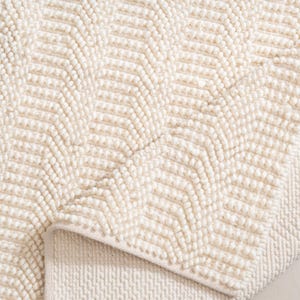 May include: Cream-colored rug with a textured, chevron pattern. The rug's design features rows of raised, rounded elements, creating a tactile surface. The edge of the rug is visible, showing a contrasting, flat border.