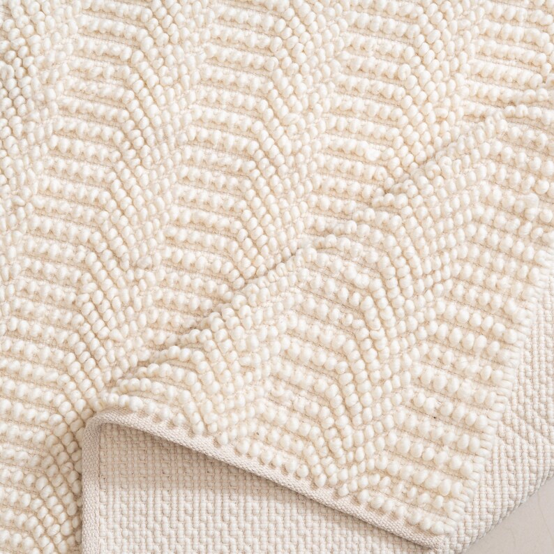 May include: Cream-colored rug with a textured, chevron pattern. The rug's design features rows of raised, rounded elements, creating a tactile surface. The edge of the rug is visible, showing a contrasting, flat border.