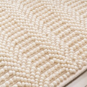 May include: Cream-colored bath mat with a textured, raised dot pattern. The soft, plush surface features a chevron design created by rows of small, rounded tufts. The edges are finished with a simple, woven border, providing a clean, finished look.