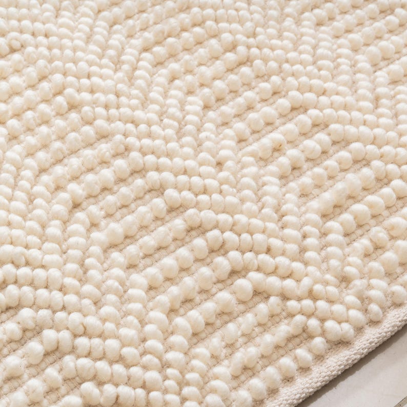May include: Cream-colored bath mat with a textured, raised dot pattern. The soft, plush surface features a chevron design created by rows of small, rounded tufts. The edges are finished with a simple, woven border, providing a clean, finished look.