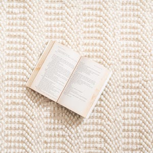 May include: An open book with aged pages rests on a cream-colored, textured rug. The rug features a repeating geometric pattern of raised, rounded elements, creating a soft, inviting surface. The book's pages display printed text.