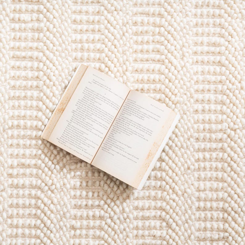 May include: An open book with aged pages rests on a cream-colored, textured rug. The rug features a repeating geometric pattern of raised, rounded elements, creating a soft, inviting surface. The book's pages display printed text.