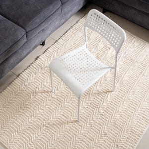 May include: A white, modern chair with a perforated back and seat sits on a cream-colored, textured rug. The rug has a geometric pattern and is placed on a light-colored floor. A gray sofa is partially visible in the background.