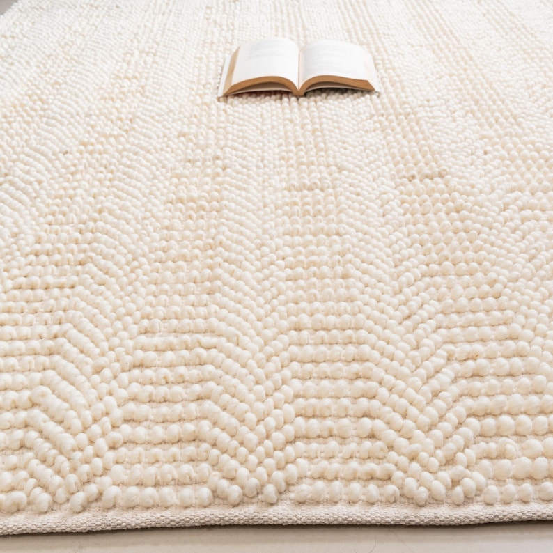 May include: Cream-colored, textured rug with a chevron pattern. The rug has a thick, looped pile and a soft, inviting appearance. An open book rests on the rug, suggesting a cozy reading nook. The rug's neutral color makes it versatile for various interior styles.