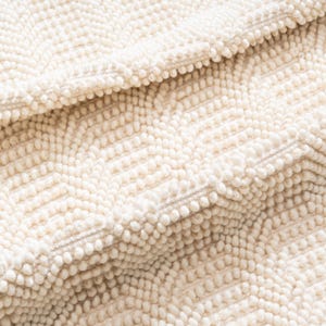 May include: Close-up of a cream-colored, textured rug with a chevron pattern. The rug has a dense, looped pile, creating a soft, tactile surface. The design features rows of small, raised loops, adding visual interest and dimension to the textile.