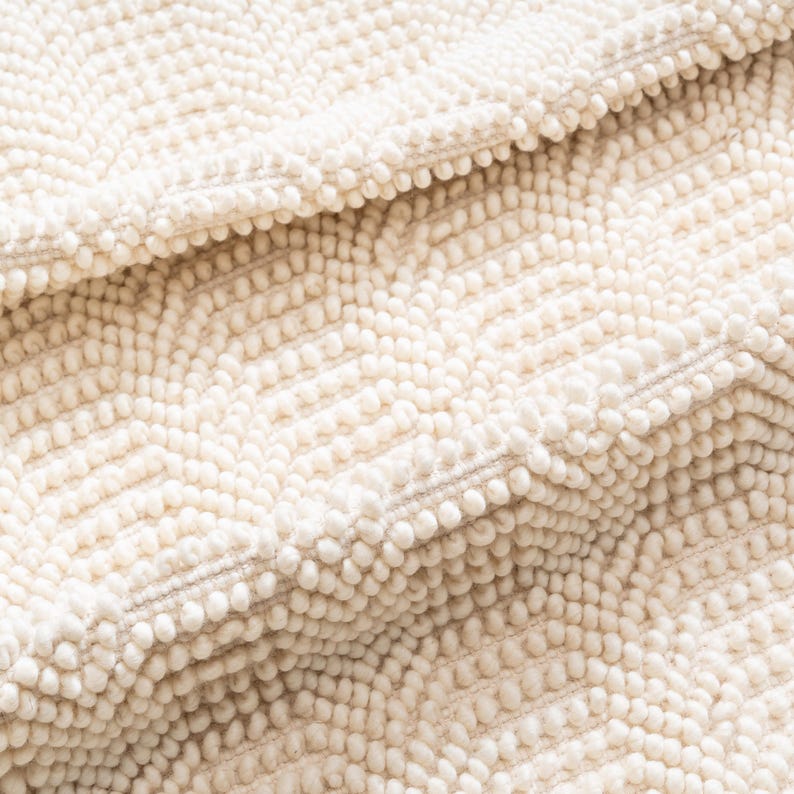 May include: Close-up of a cream-colored, textured rug with a chevron pattern. The rug has a dense, looped pile, creating a soft, tactile surface. The design features rows of small, raised loops, adding visual interest and dimension to the textile.
