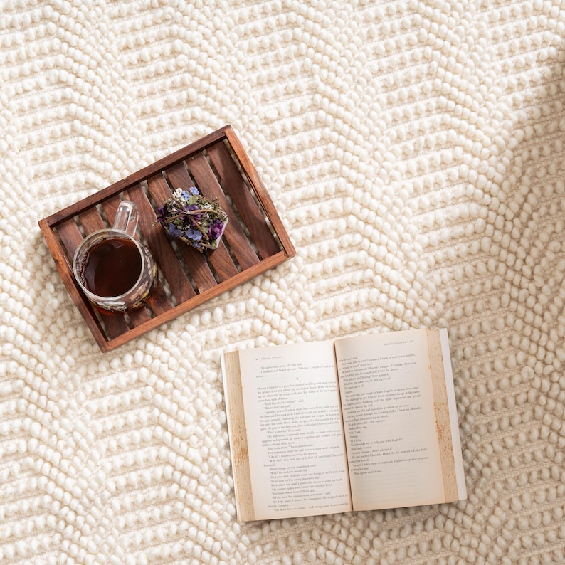 May include: A cozy scene featuring a cream-colored, textured rug with a chevron pattern. A wooden tray holds a cup of tea and dried flowers. An open book rests nearby, suggesting relaxation and leisure.