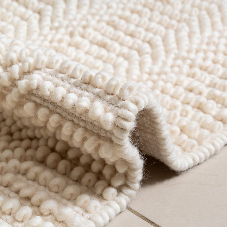 May include: Cream-colored, textured rug with a looped, bobble-like surface. The rug features a geometric pattern and a folded edge, showcasing its thickness and texture. The neutral color and tactile design suggest a cozy, home decor item.