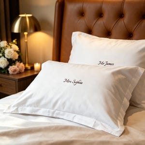 Puede incluir: Dos fundas de almohada rectangulares blancas con borde de volante. La funda delantera presenta el texto bordado Mrs Sophia en una fuente cursiva oscura, mientras que la funda trasera muestra el texto bordado Mr James en la misma fuente oscura.