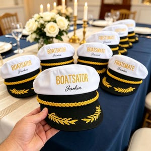 Op de afbeelding: Een wit met zwarte kapiteinspet met goudkleurig borduurwerk met de tekst BOATSATOR of FIRSTSMATE, een gouden gevlochten koord aan de voorzijde en een gouden lauwerkransmotief op de zwarte klep, vastgehouden boven een gedekte tafel.