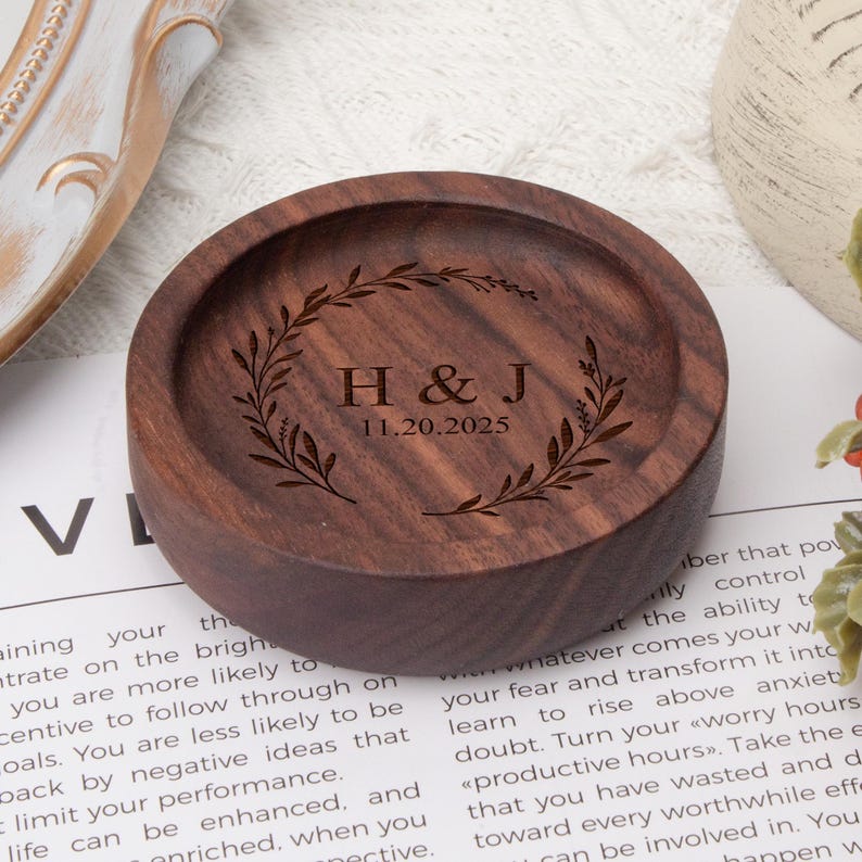 May include: A round wooden ring holder with a decorative engraved wreath and the initials "H&J" and the date "11.20.2025".