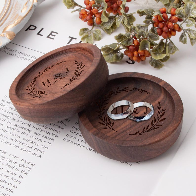 May include: Two wooden ring boxes with engraved leafy wreaths and the initials "H & J" and the date "11.20.2025". The boxes are stacked on top of each other.