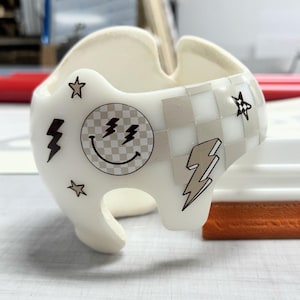 May include: A white helmet with a checkered pattern and various stickers, including a smiley face with lightning bolts, stars, and lightning bolts. The helmet is next to a white and orange squeegee.