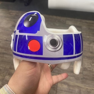 May include: A white helmet with a blue and white R2-D2 design. The helmet has a red circle, a black circle, and a blue rectangle on the front.