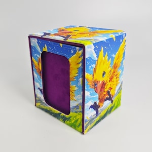 May include: A square deck box featuring a vibrant illustration of a yellow bird-like creature with orange accents against a blue sky and green field. The box has a purple interior lining and a rectangular cutout on the front panel for viewing.