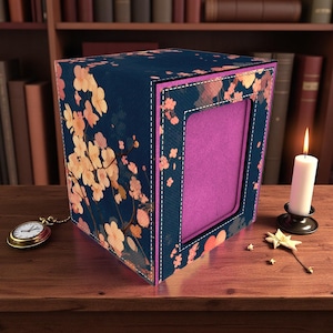 Puede incluir: Una caja decorativa con un estampado floral en tonos melocotón y blanco sobre un fondo azul marino. La caja tiene un interior rosa y un borde cosido. Una vela encendida y un reloj de bolsillo están sobre la superficie de madera.